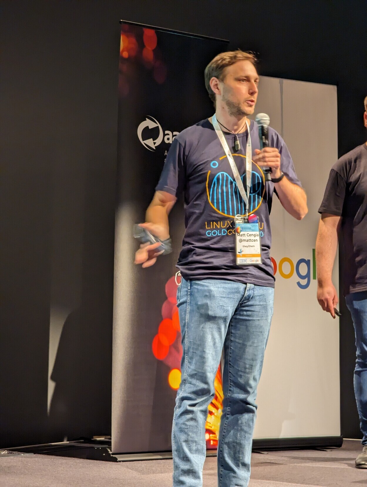 Matt, a masc-presenting person with a short beard, standing on stage
in jeans and a T-shirt, holding a fist microphone and gesticulating with a
presenter remote, while talking to an audience. They're wearing a lanyard with
some LGBTQ+ badges and a name tag. In the background are banners branded Google
and aarnet.